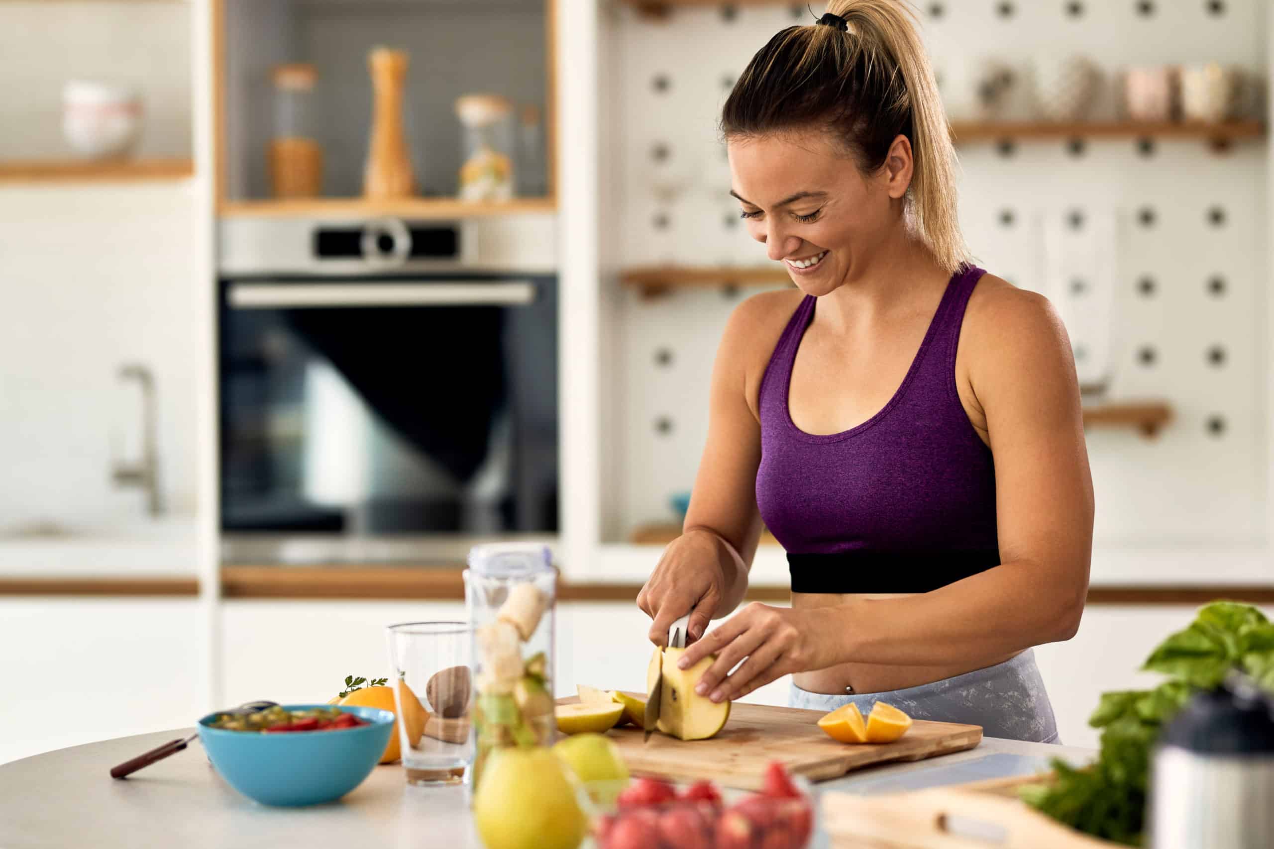happy-athletic-woman-cutting-fruit-while-preparing-healthy-meal-kitchen-scaled