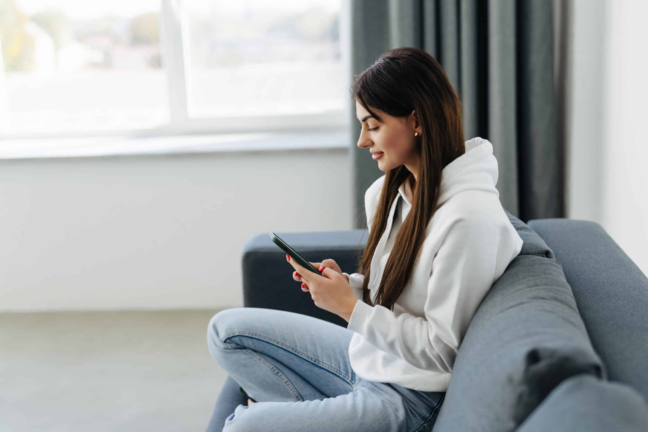 smiling-young-woman-using-phone-sitting-couch-home-looking-smartphone-screen-beautiful-female-chatting-social-network-shopping-online-spending-leisure-time-with-mobile-device-scaled