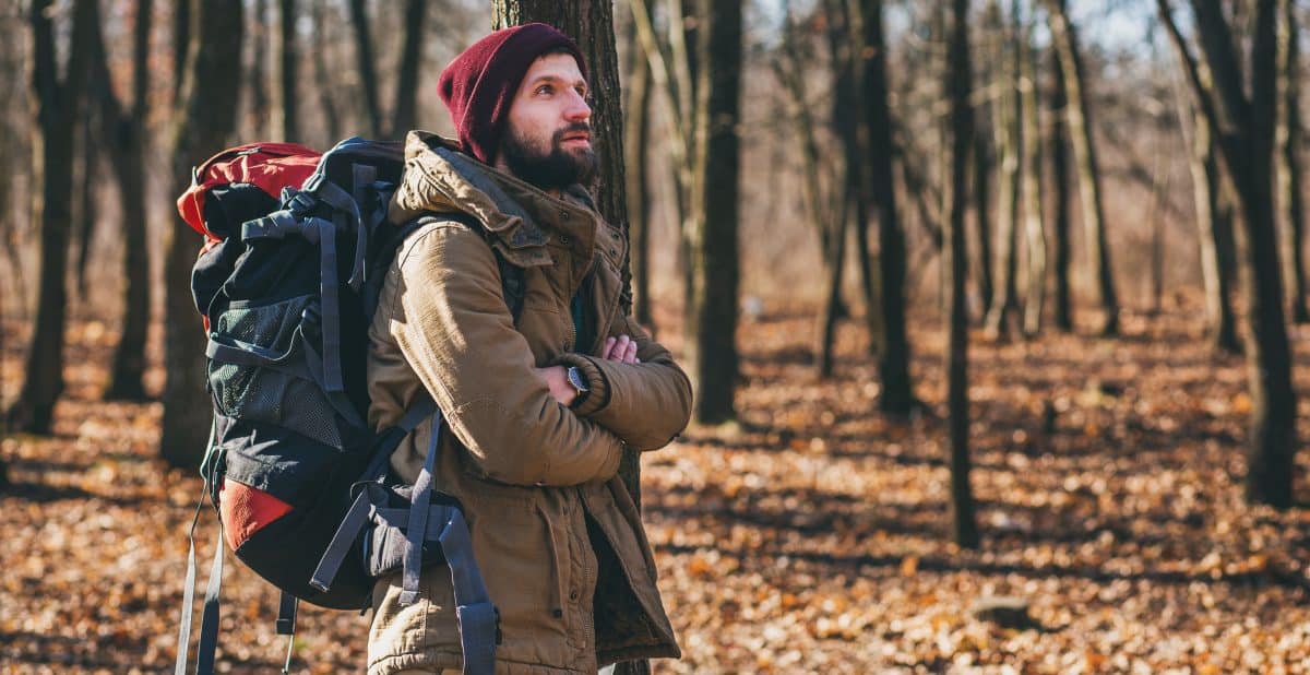 young-hipster-man-traveling-with-backpack-autumn-forest-wearing-warm-jacket-hat-1-e1637333607360